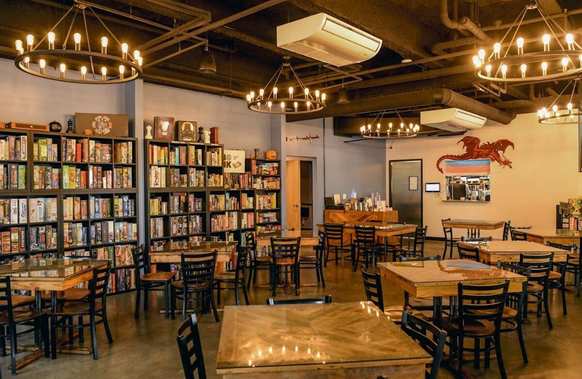 Discover The Castle, your ultimate board game café experience! Dive into a warm and welcoming atmosphere with shelves bursting with games, inviting wooden tables and chairs, and charming circular chandeliers that light up the room. Enhance your adventure with the playful red dragon decoration on one wall, adding a whimsical twist to this enchanting haven. Perfect for game enthusiasts and casual visitors alike!.