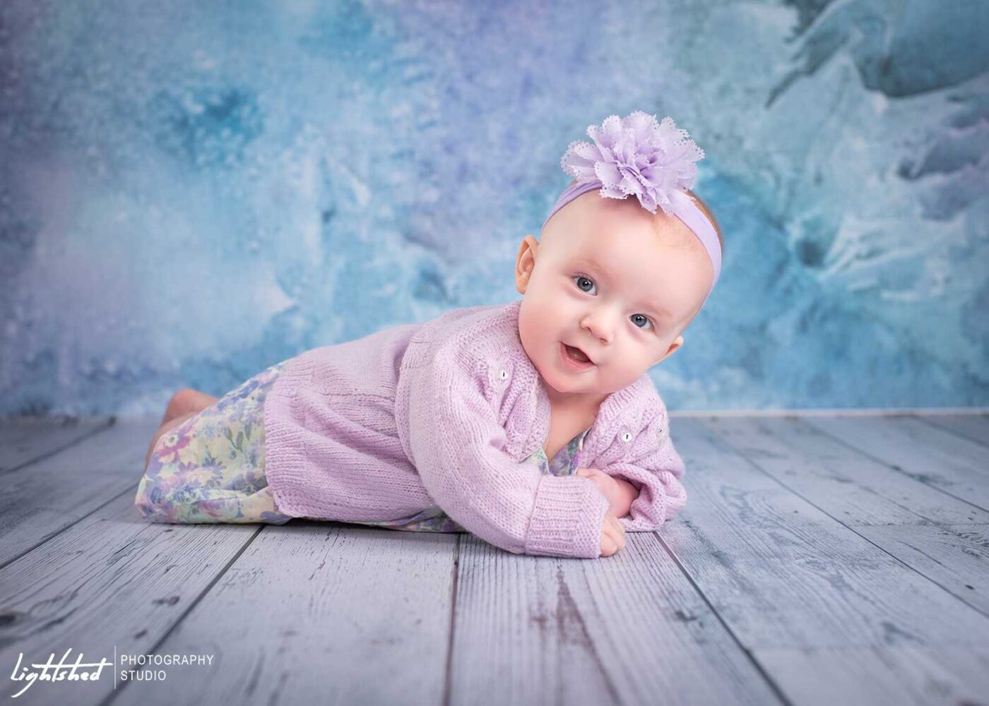 A baby wearing a lavender headband with a flower and a light purple sweater lies on a wooden floor with a blue and purple textured backdrop. Captured by Kendra St. John, the baby looks at the camera with a slight smile.