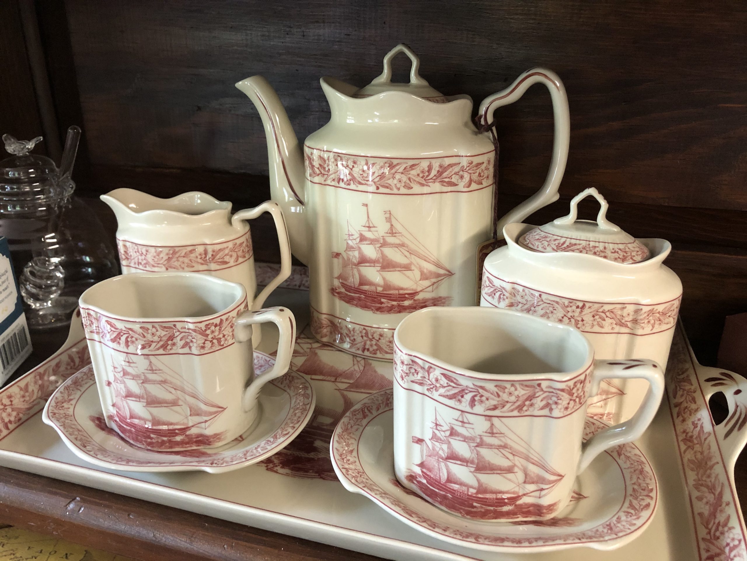 A ceramic tea set with a red ship design, perfect for a virtual tea party or fans of the House of the Seven Gables, includes a teapot, creamer, sugar bowl, two cups with saucers, and a rectangular tray on a wooden shelf.