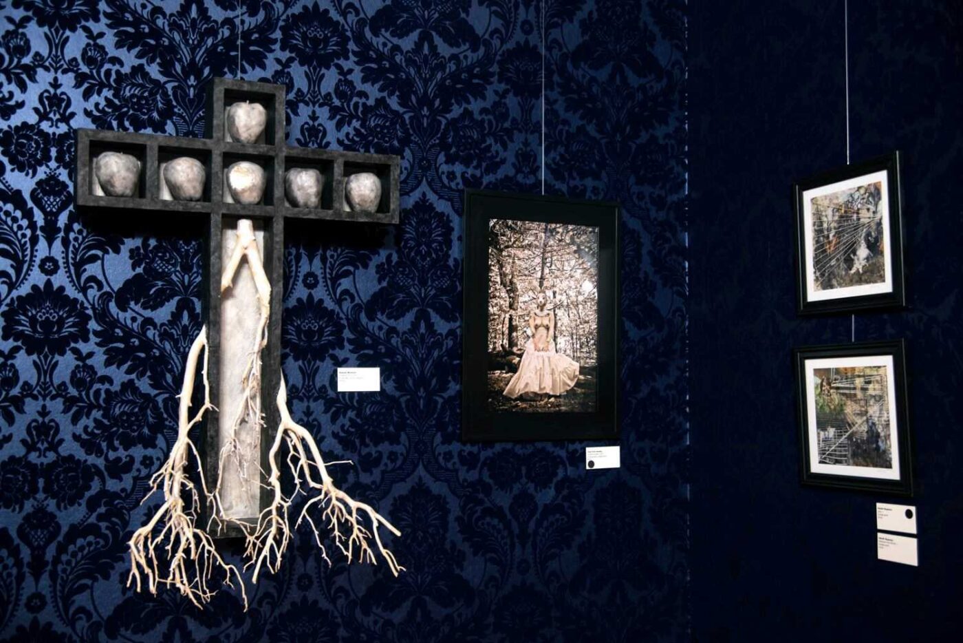A dark blue, patterned wall at Salem Art Gallery displays three artworks: a cross with roots and silver objects, a central photograph of a woman in white in a forest, and two framed mixed media pieces. Each artwork has a small label.