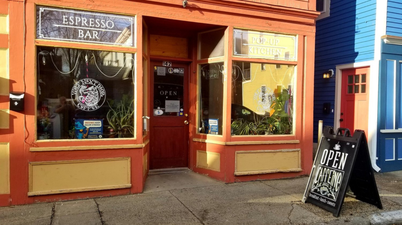 Discover a charming spot where coffee enthusiasts and food lovers meet: the bright red and orange storefront labeled "Espresso Bar" and "Pop-Up Kitchen." An inviting "Open" sign welcomes you at the entrance, while a sandwich board announces: "Open Cafeino by Wolf Next Door Coffee.