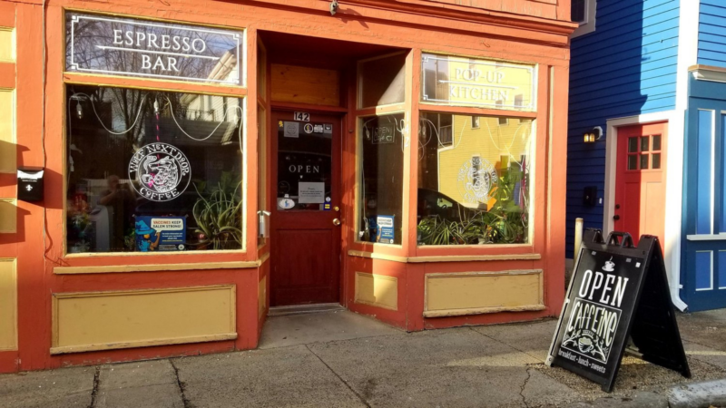 Discover a charming spot where coffee enthusiasts and food lovers meet: the bright red and orange storefront labeled "Espresso Bar" and "Pop-Up Kitchen." An inviting "Open" sign welcomes you at the entrance, while a sandwich board announces: "Open Cafeino by Wolf Next Door Coffee.