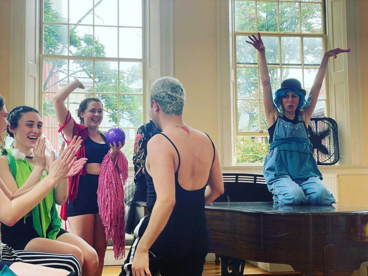 Four people are in a bright room with large windows. Three stand and cheer, one holding a purple ball and pink fringe. Another sits on a piano, arms raised in a blue outfit and hat—like DREAM LOVE ESCAPE’s Free Public Performances in Salem MA.