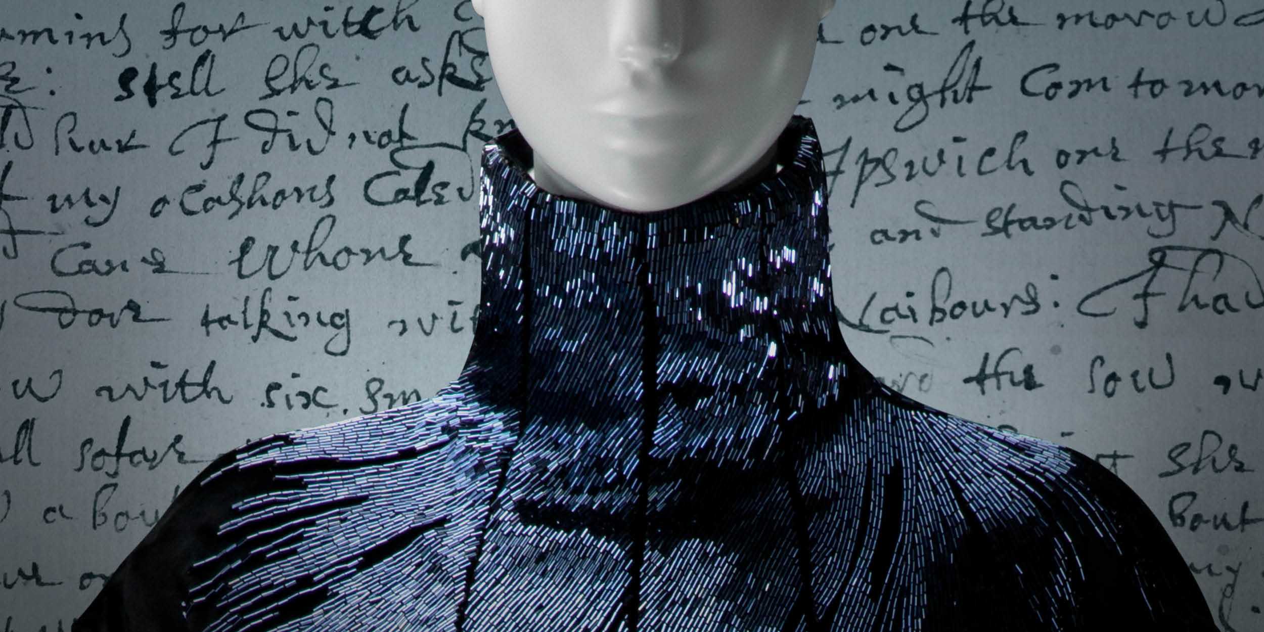 A mannequin dressed in a high-neck, black garment with shimmering, silver sequin embellishments stands before a backdrop of handwritten script, evoking the history and spirit of Reckoning and Reclaiming from the era of the Salem Witch Trials.
