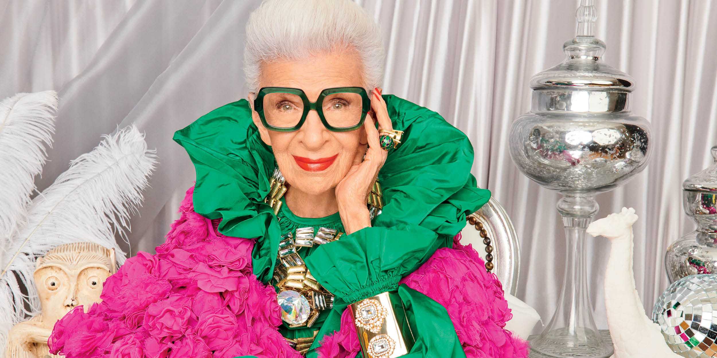 Channeling Iris Apfel, an older woman with white hair, oversized black glasses, and bright red lipstick wears a vibrant green and pink ruffled outfit with large jewelry, celebrating in front of silver decor and white feathers—perfect for a century celebration.