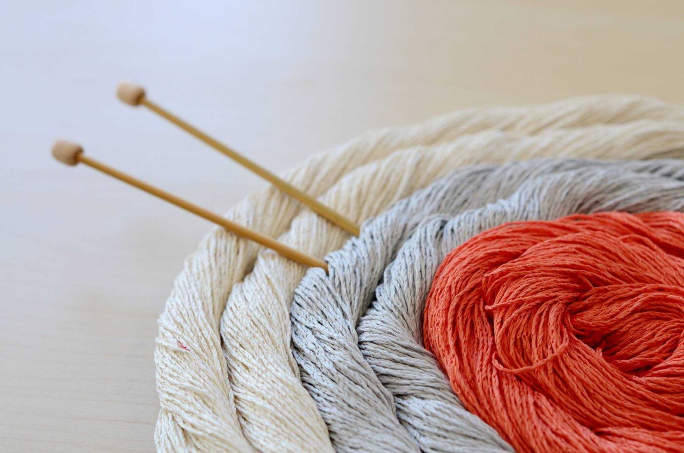 Close-up of three coils of yarn from Circle of Stitches in cream, gray, and orange hues arranged in a spiral, with two wooden knitting needles resting across the coils—perfect for crafting your own origin stories.