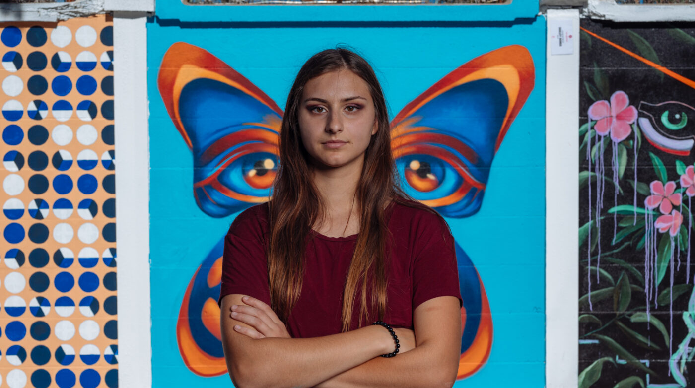 A person with long hair stands confidently, arms crossed, in front of a vivid mural showcasing butterfly wings. This striking creation highlights the dynamic creativity of Flourish Artistic Services.
