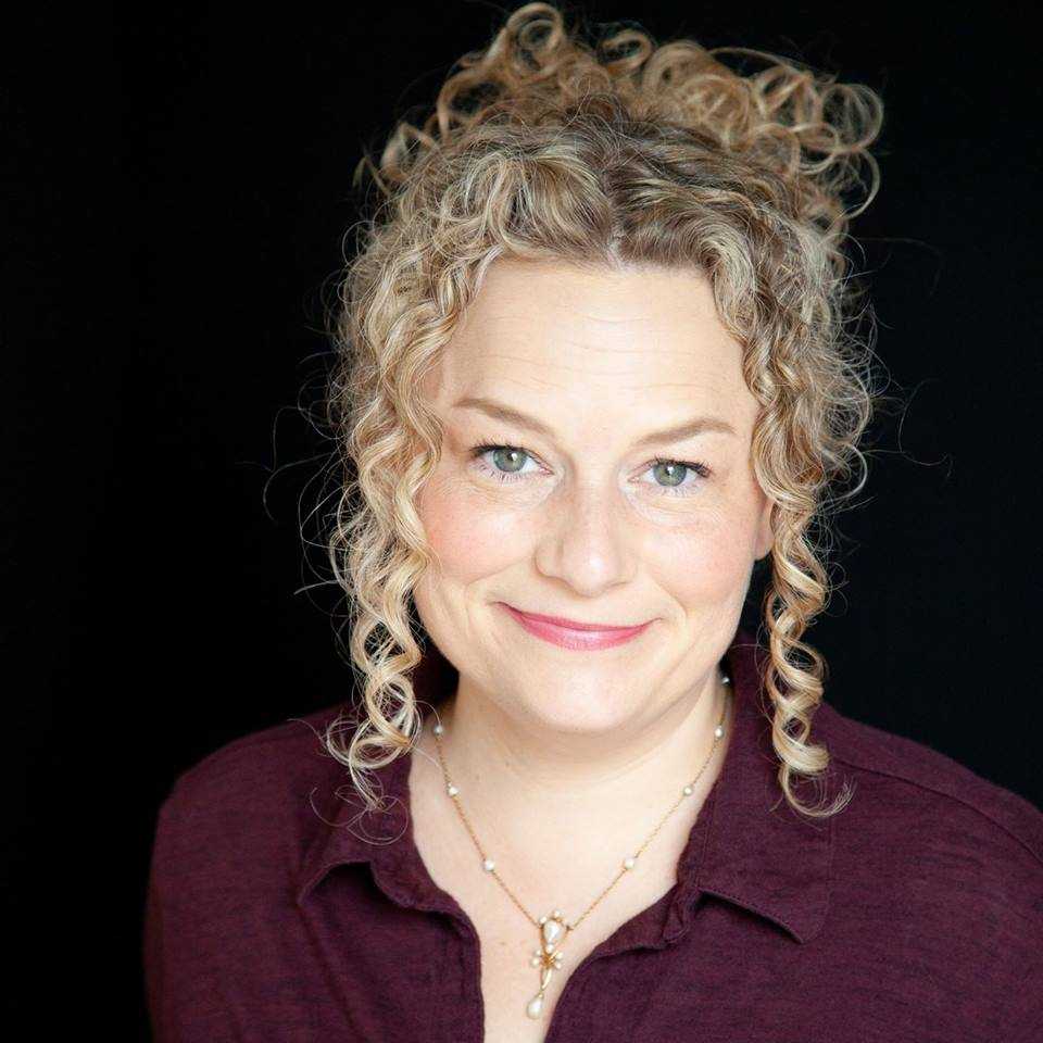 A woman with curly blonde hair pulled up, wearing a maroon button-up shirt and a pearl necklace, smiles slightly while looking at the camera. The black background suggests a Live Theatre setting, reminiscent of an ORLANDO production at Salem State.