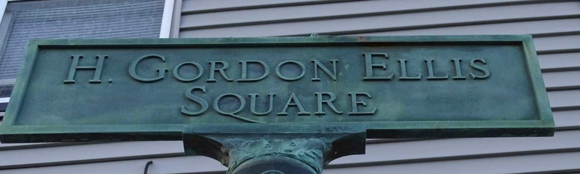 A metal street sign reads "H. Gordon Ellis Square" in capital letters, marking one of Beverly's Veterans Squares, with a building featuring horizontal siding and a window in the background.