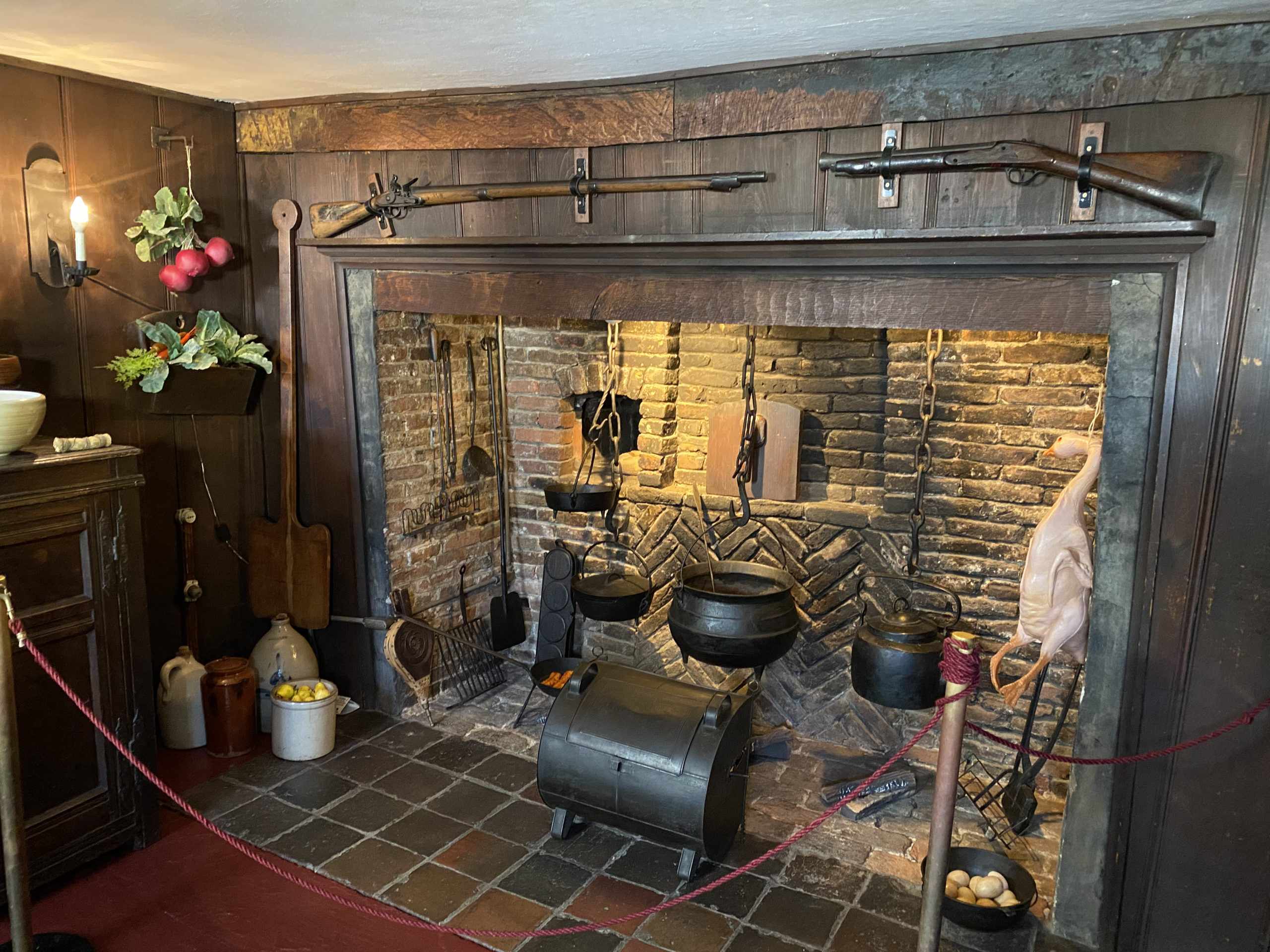 A colonial-era kitchen hearth display with cast iron pots, cooking tools, and two muskets mounted above the brick fireplace captures New England flavor. Ceramic jugs, vegetables, a plucked fowl, and a modest cake are staged nearby behind a rope barrier.