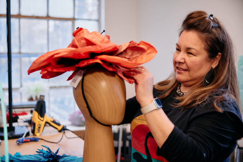 In a Grace & Diggs workshop, you’ll find creativity in full bloom. Picture this: a woman perfecting the fit of an oversized red paper flower hat on a mannequin, while her workspace buzzes with energy from nearby glue guns and art supplies. Dive into the vibrant world of DIY fashion and let your imagination run wild!.