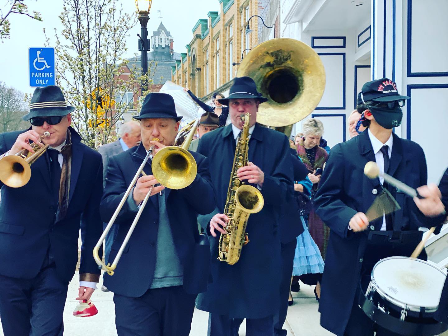four men in black suits and black hats play musical instruments on the street