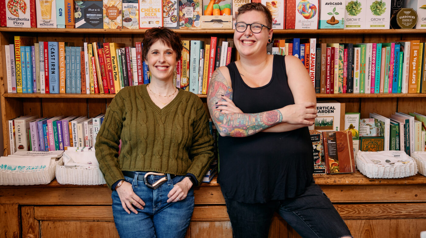 In front of a packed bookshelf, two individuals express their unique styles. One sports a green sweater with jeans for a comfy, casual look. The other person rocks a black tank top that highlights their vibrant tattoos, adding an artistic flair to the scene.
