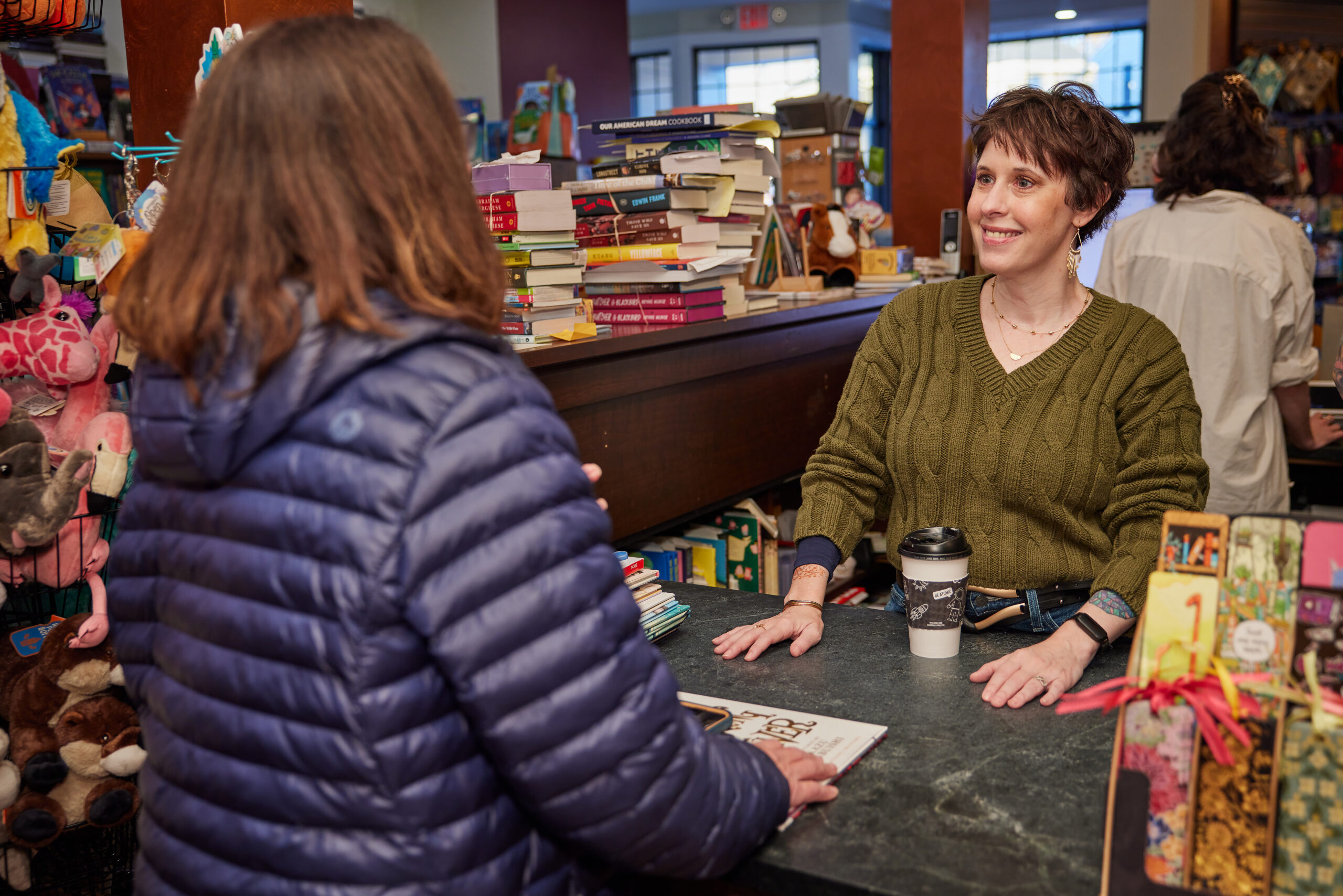 A friendly individual in a green sweater chats with a customer in a blue jacket at the bookstore counter, surrounded by an array of books and cute stuffed animals.