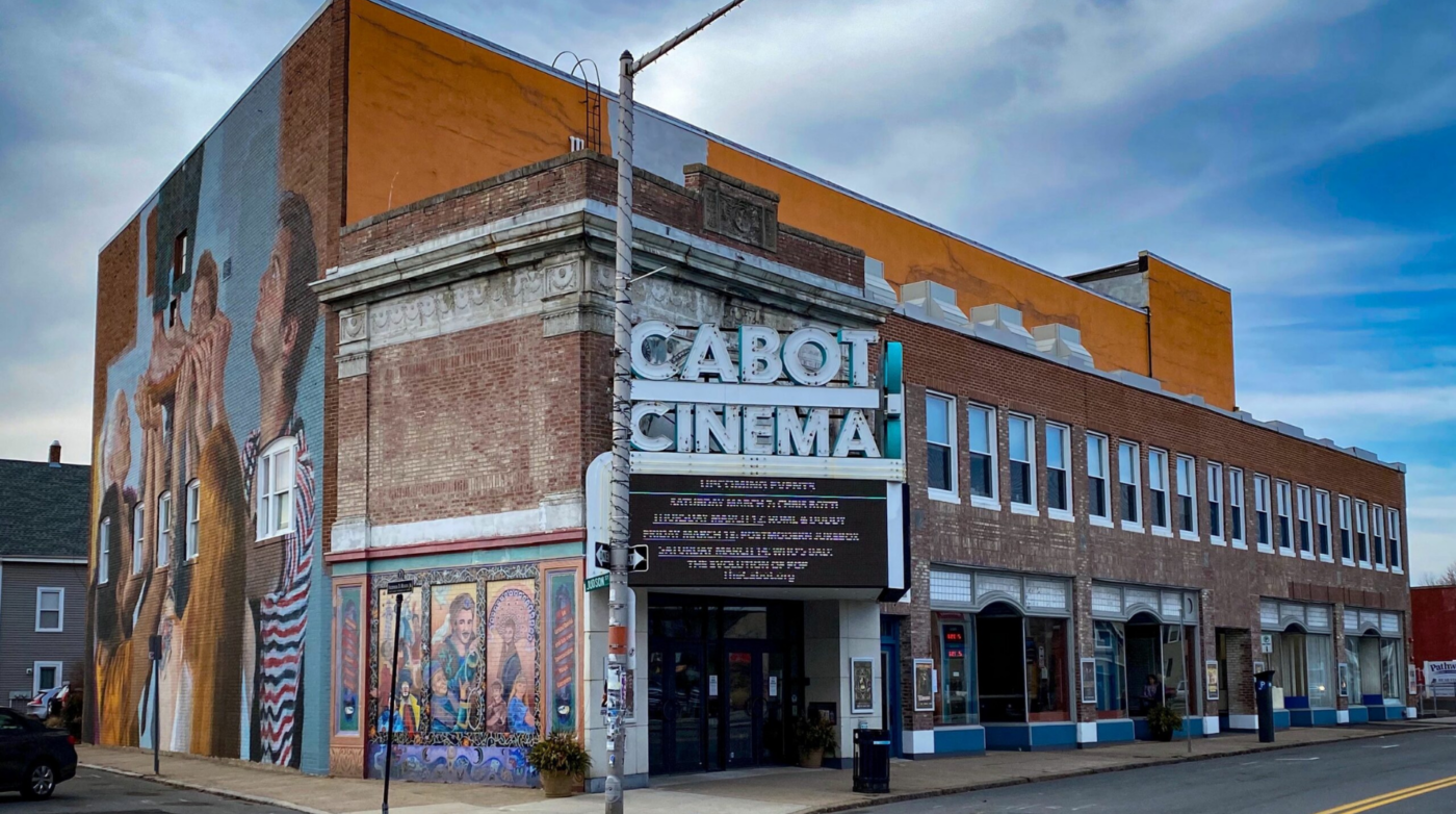 Discover The Cabot Cinema, a charming historic brick building that captures attention with its stunning side mural. Its timeless marquee showcases the latest film listings, set against the beauty of a partly cloudy sky.