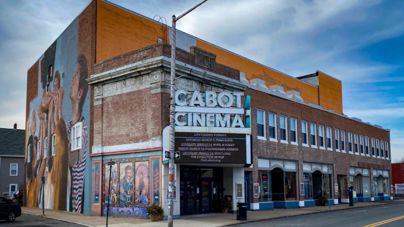 Discover The Cabot Cinema, a charming historic brick building that captures attention with its stunning side mural. Its timeless marquee showcases the latest film listings, set against the beauty of a partly cloudy sky.