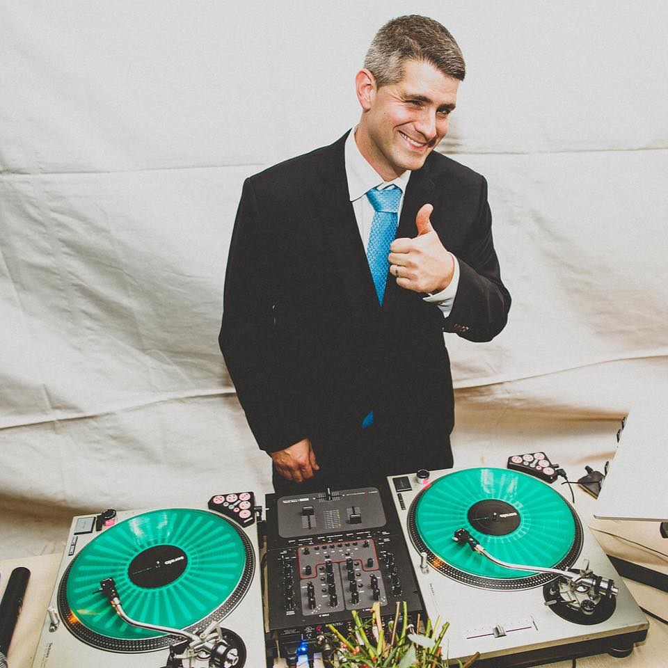 a white man with short grey hair in a black suit and blue tie stands in front of a dj table. He is giving a thumbs up to the camera.
