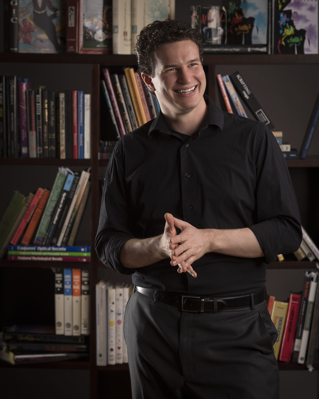 a white man with short black hair stands in front of a bookshelf. He is wearing a black button up shirt and black pants.