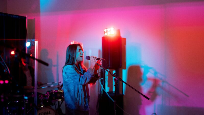 A person in a denim jacket passionately sings into a microphone on stage, while ND Productions' vibrant red and blue lights create mesmerizing shadows.