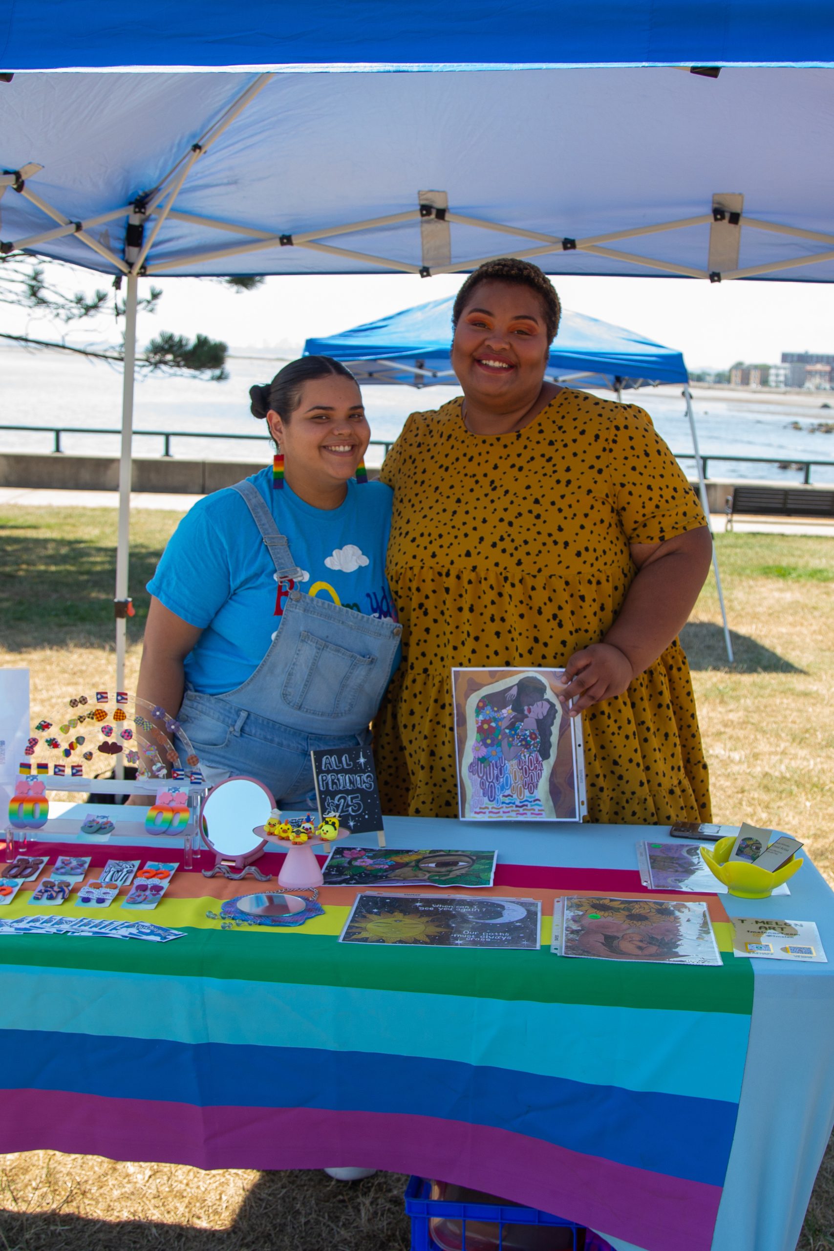 two femme people stand behind a table full of fun rainbow products. The latinx human on the left has black hair in buns and is wearing a blue t-shirt and overalls. The human on the right is black and wears a polkadotted yellow dress. 