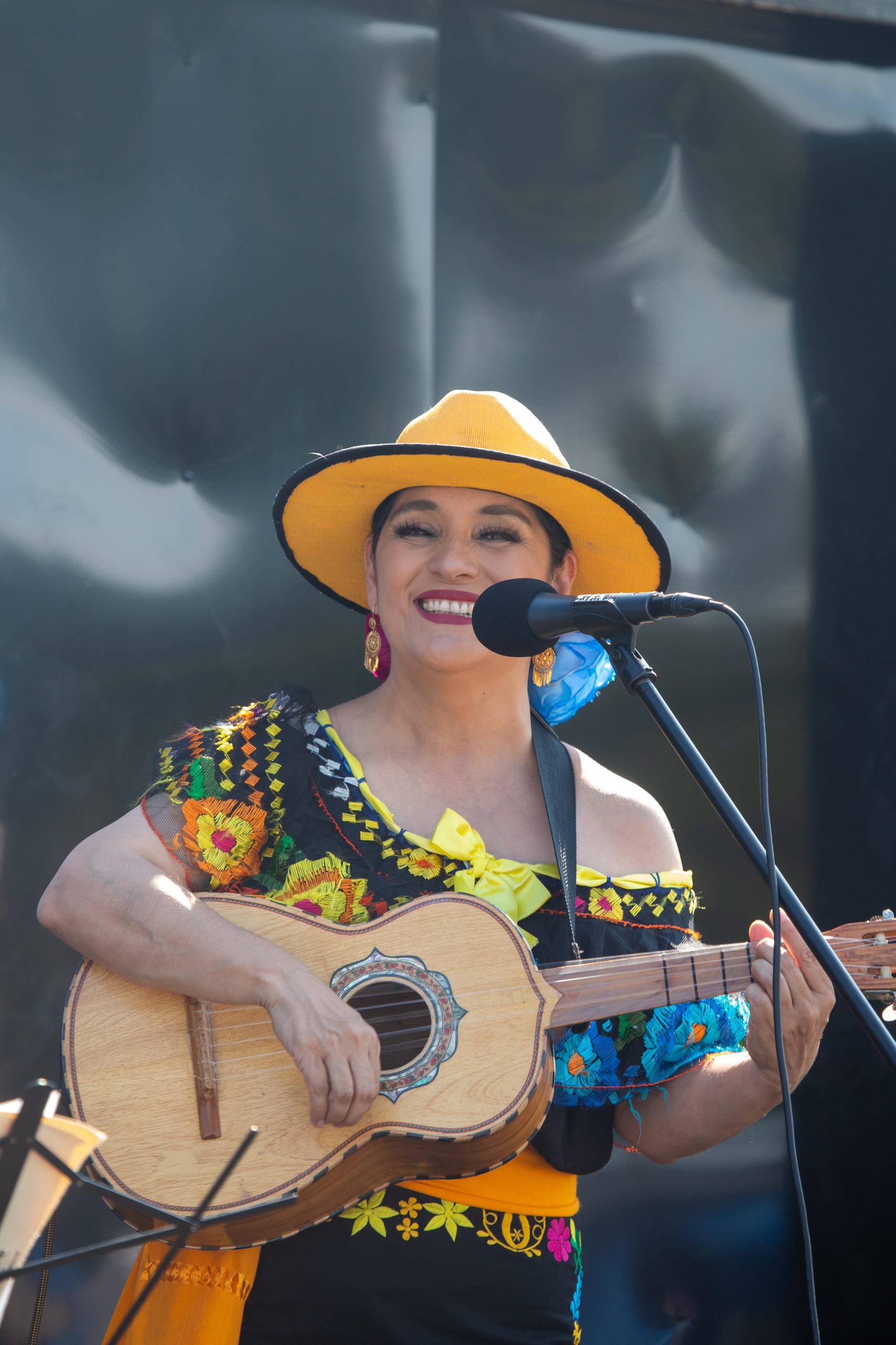 a latina woman in a yellow sombrero holds a mariachi guitar and smiles behind a microphone