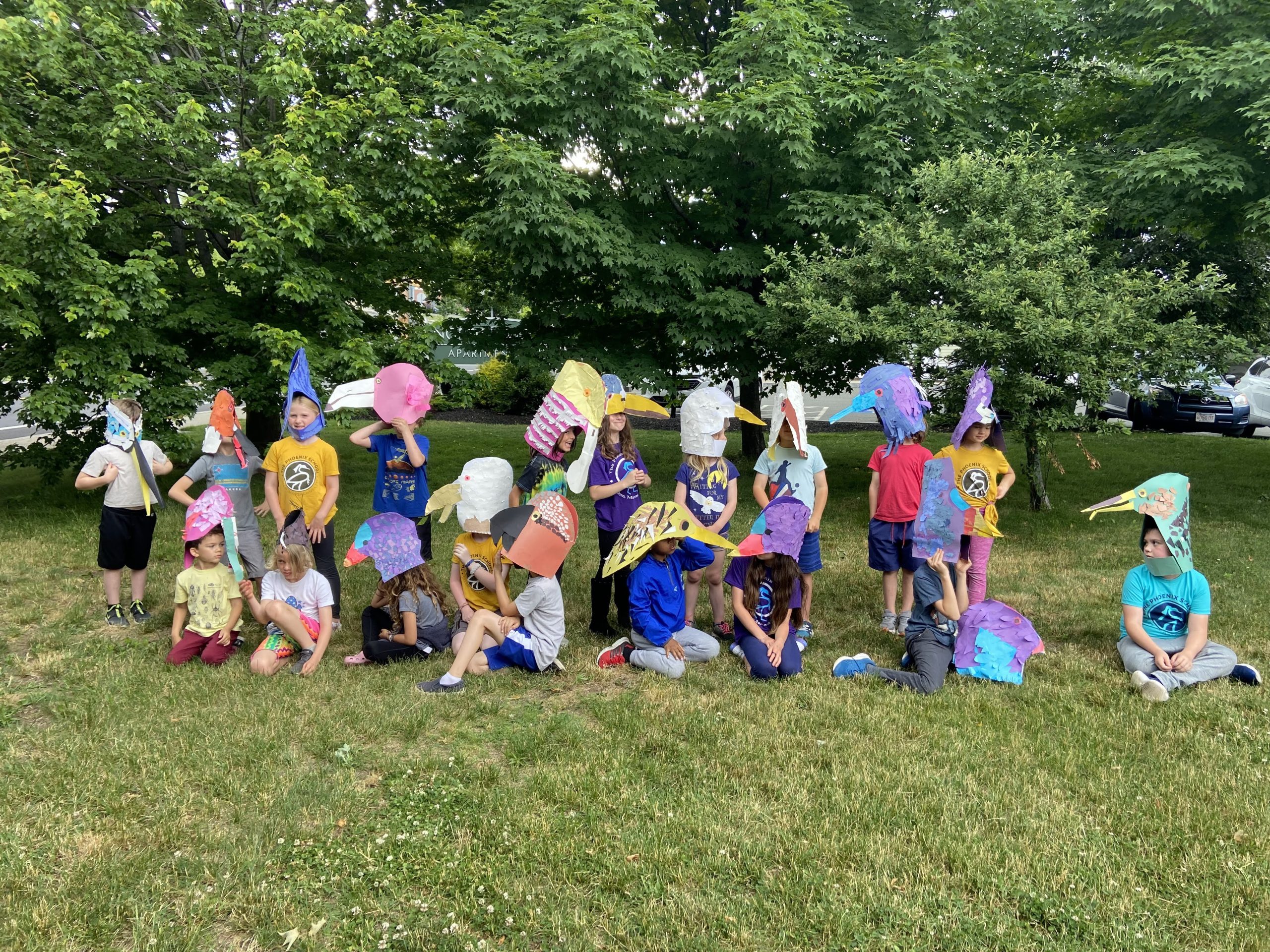 A group of children sit in the grass outside holding handmade art