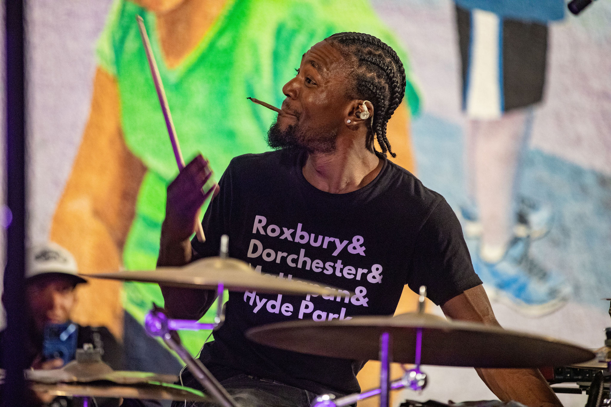 A black drummer wearing a black graphic t-shirt. Their hair is in corn rows. 
