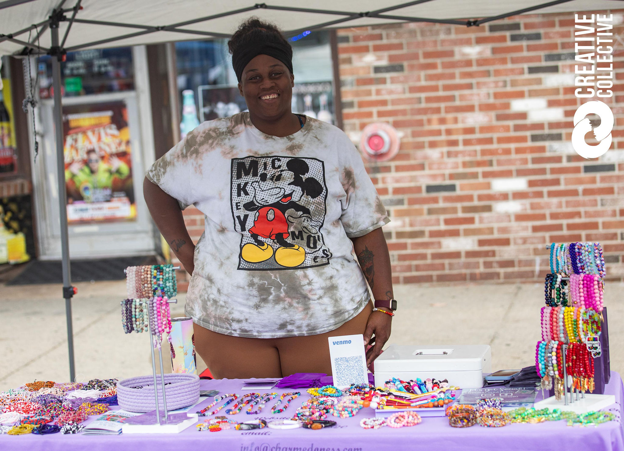 A curvy black person with short cropped hair in a mickey mouse t-shirt smiles behind her booth at the Peabody International Festival