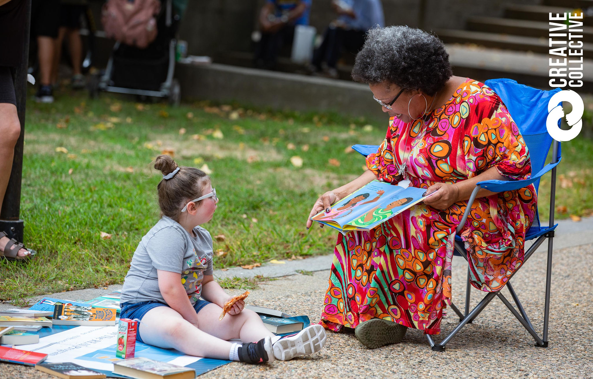 Afrolatina poet Michelle LaPoetica reads a story to a young white girl outside during the Peabody International Festival. Michelle wears a patterned red dress and the girl wears a grey t-shirt and blue shorts.