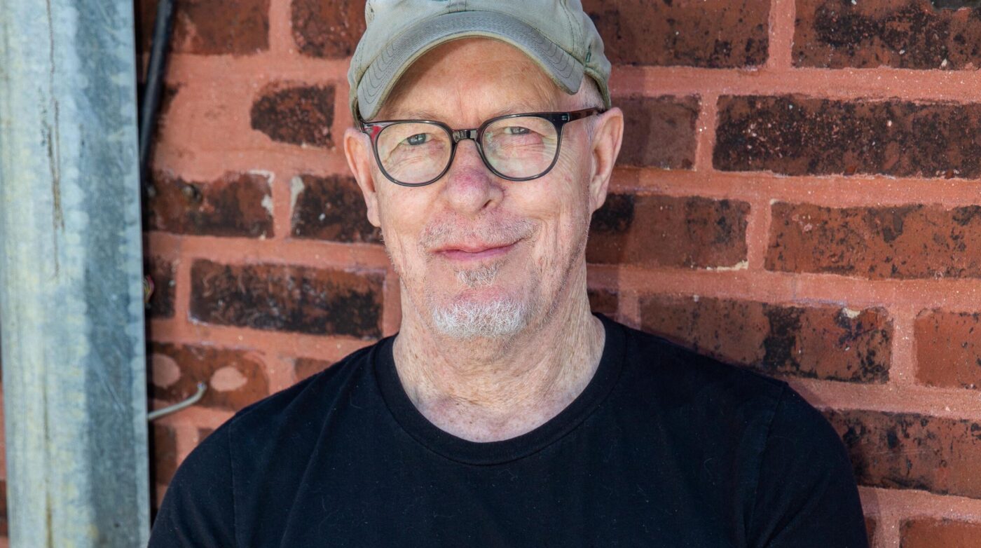 a man wearing a hat and glasses standing in front of a brick wall.