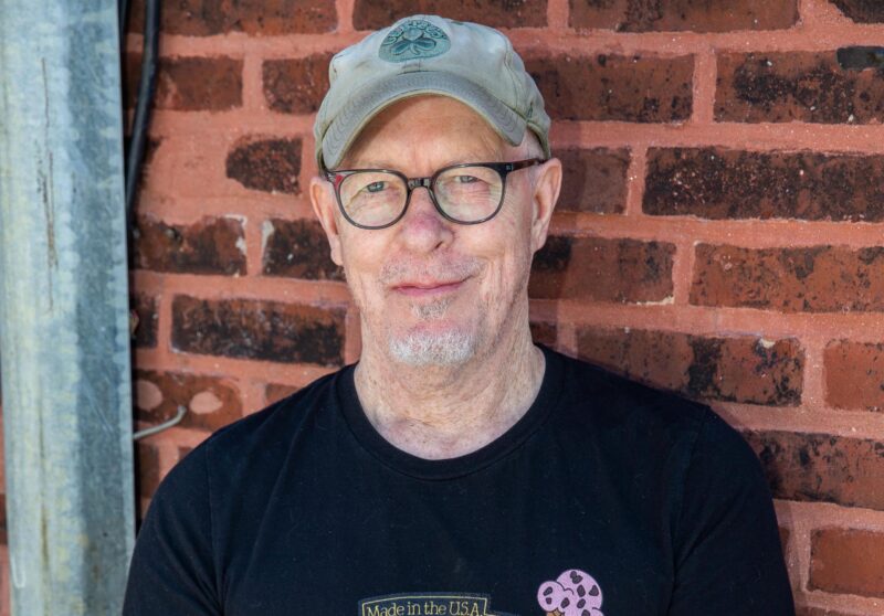 a man wearing a hat and glasses standing in front of a brick wall.