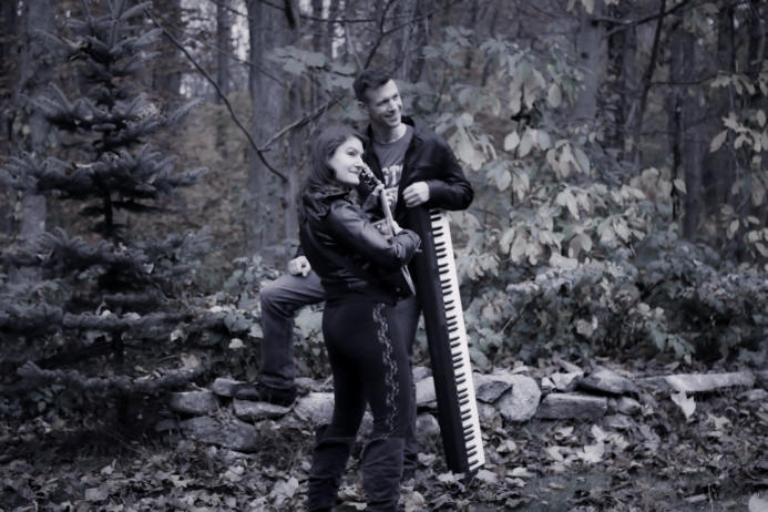 A man and woman standing next to a piano in the woods.