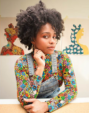 a woman in overalls with afro hair sitting at a desk.