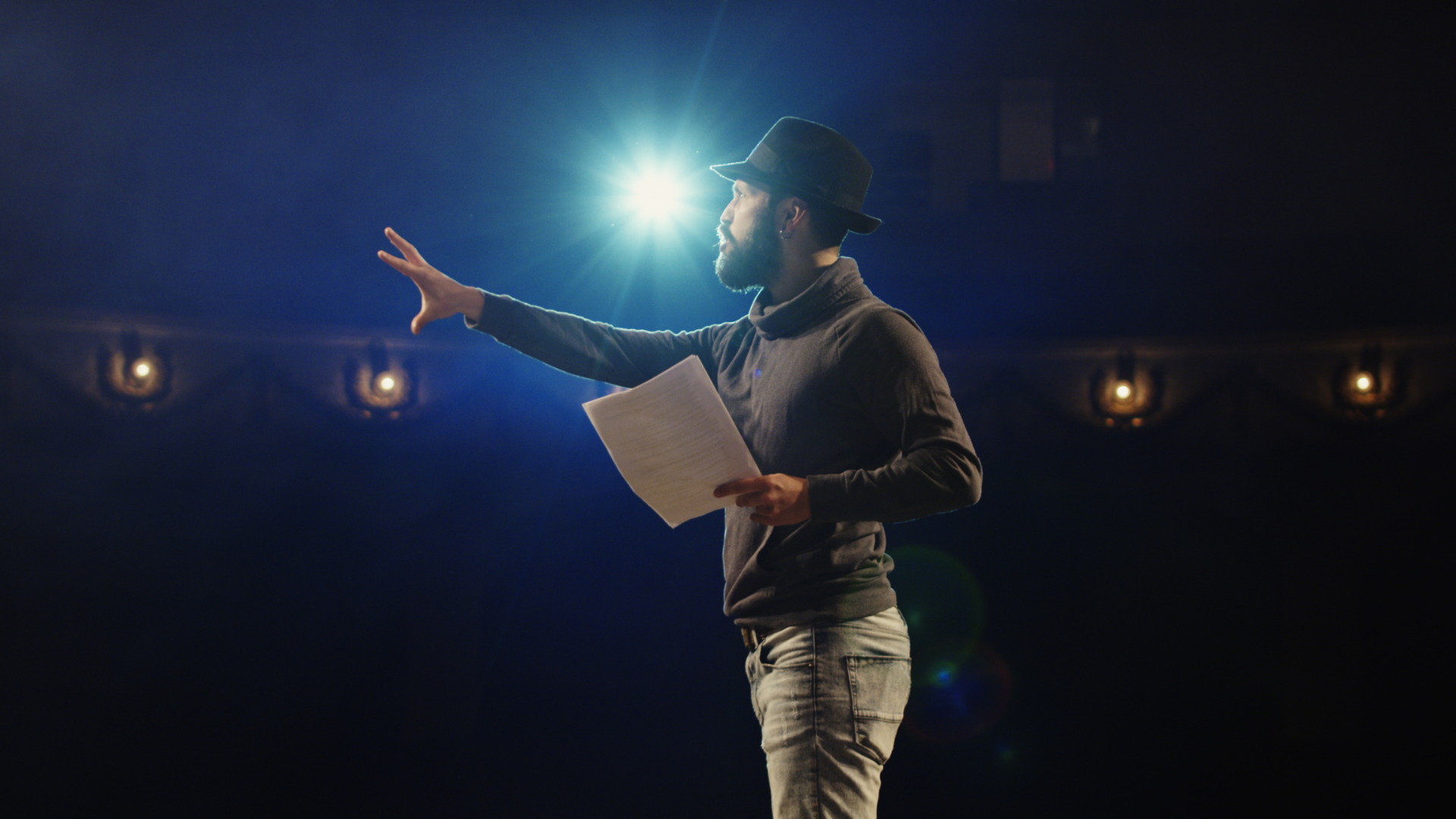 In the theatre, a man in a hat confidently holds a piece of paper in front of a spotlight.