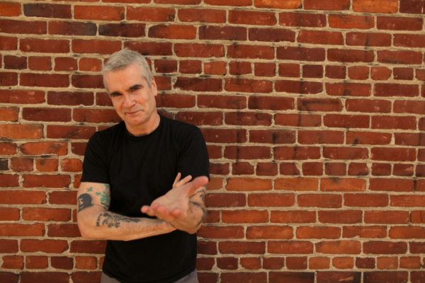 A man with tattoos leaning against a brick wall.