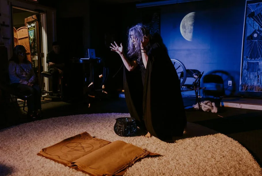 A woman in a black cloak is standing on a rug in a dark room.
