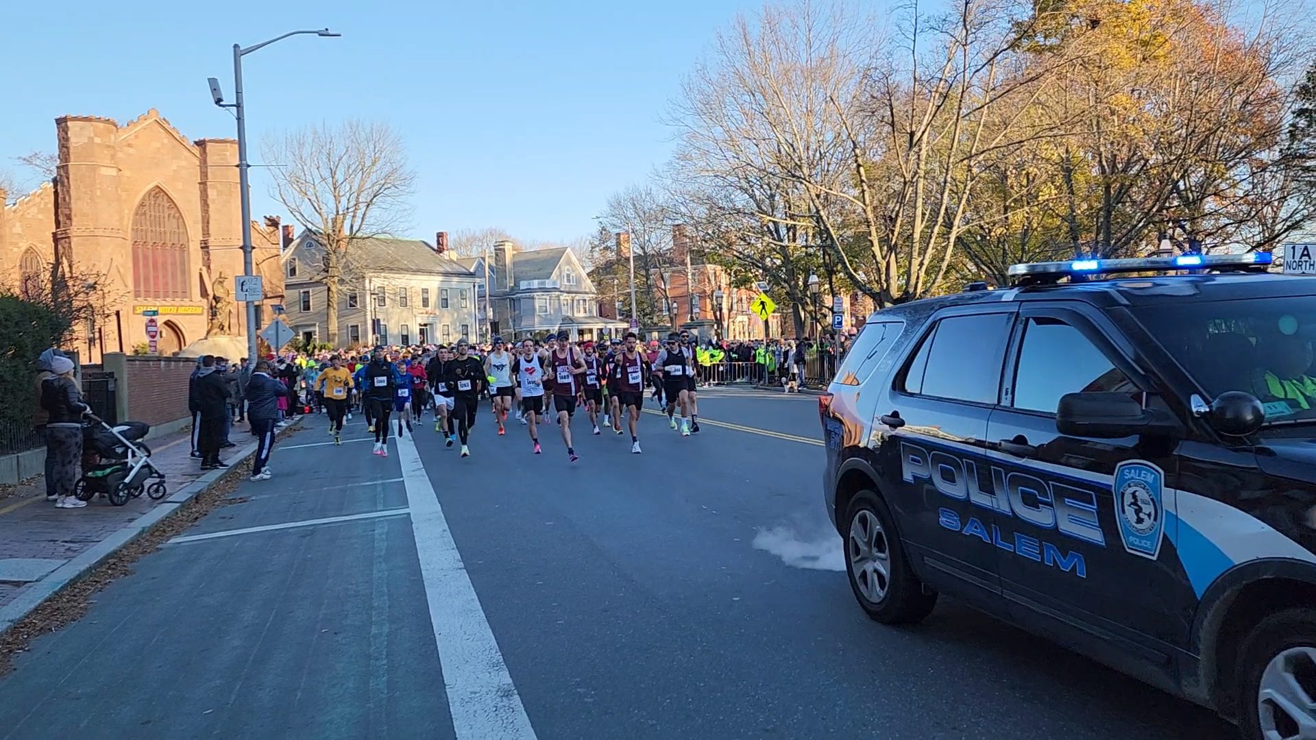 A group of runners are running down the street in front of a police car.