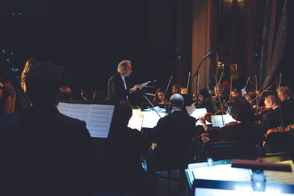 A man presenting to an eager audience with a symphony orchestra as a backdrop.