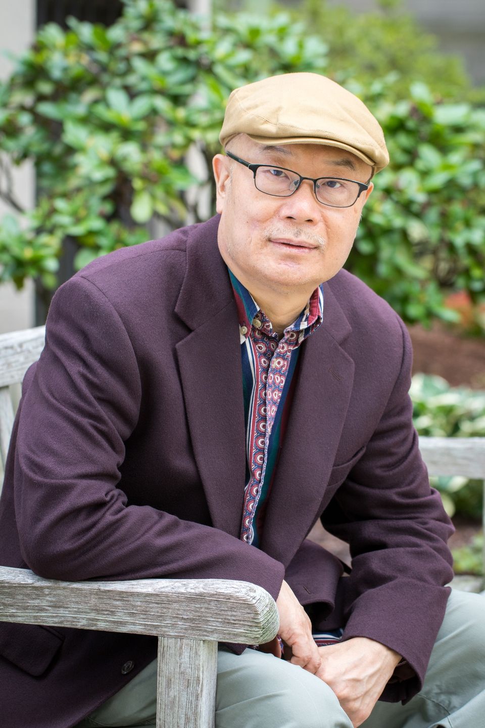 A man wearing a stylish flat cap and eyeglasses, casually relaxing on a bench surrounded by lush green plants.