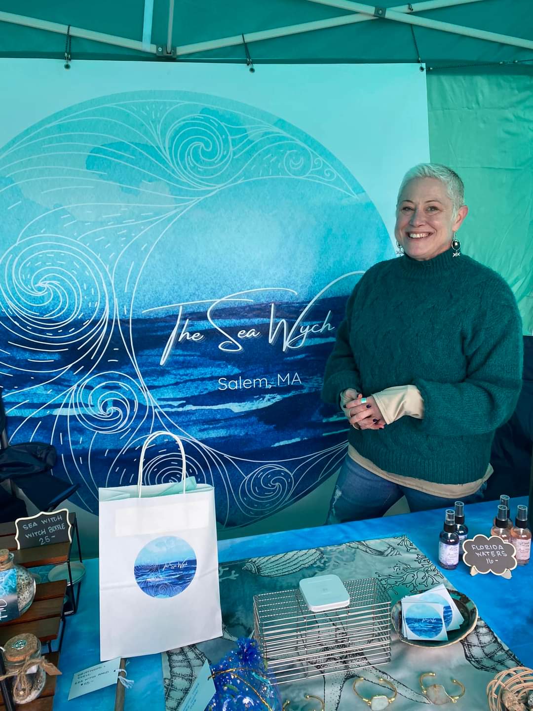 Experience the joy of shopping with a friendly seller at "The Sea Witch" in Salem, MA. Discover products displayed in a booth adorned with unique ocean-themed décor.