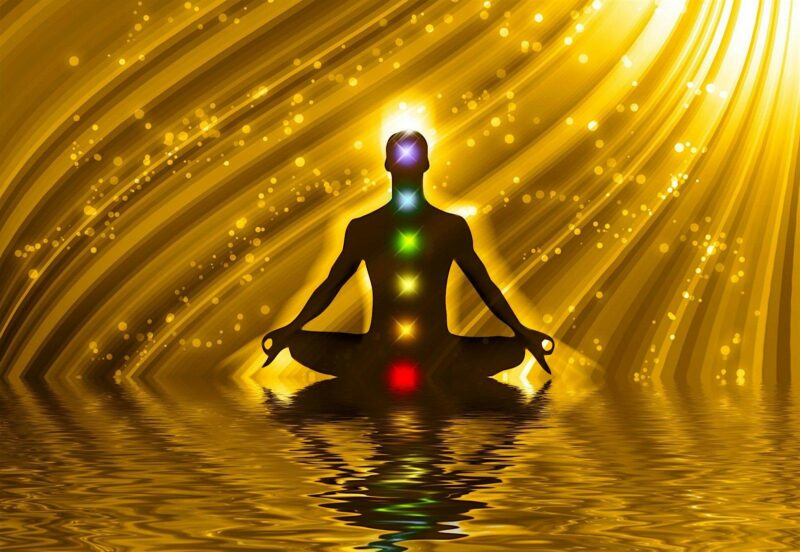 An online art piece showing a shadowy person meditating with glowing chakras on their body. This captivating image is placed over a shimmering gold background where beams of light meet towards the horizon.