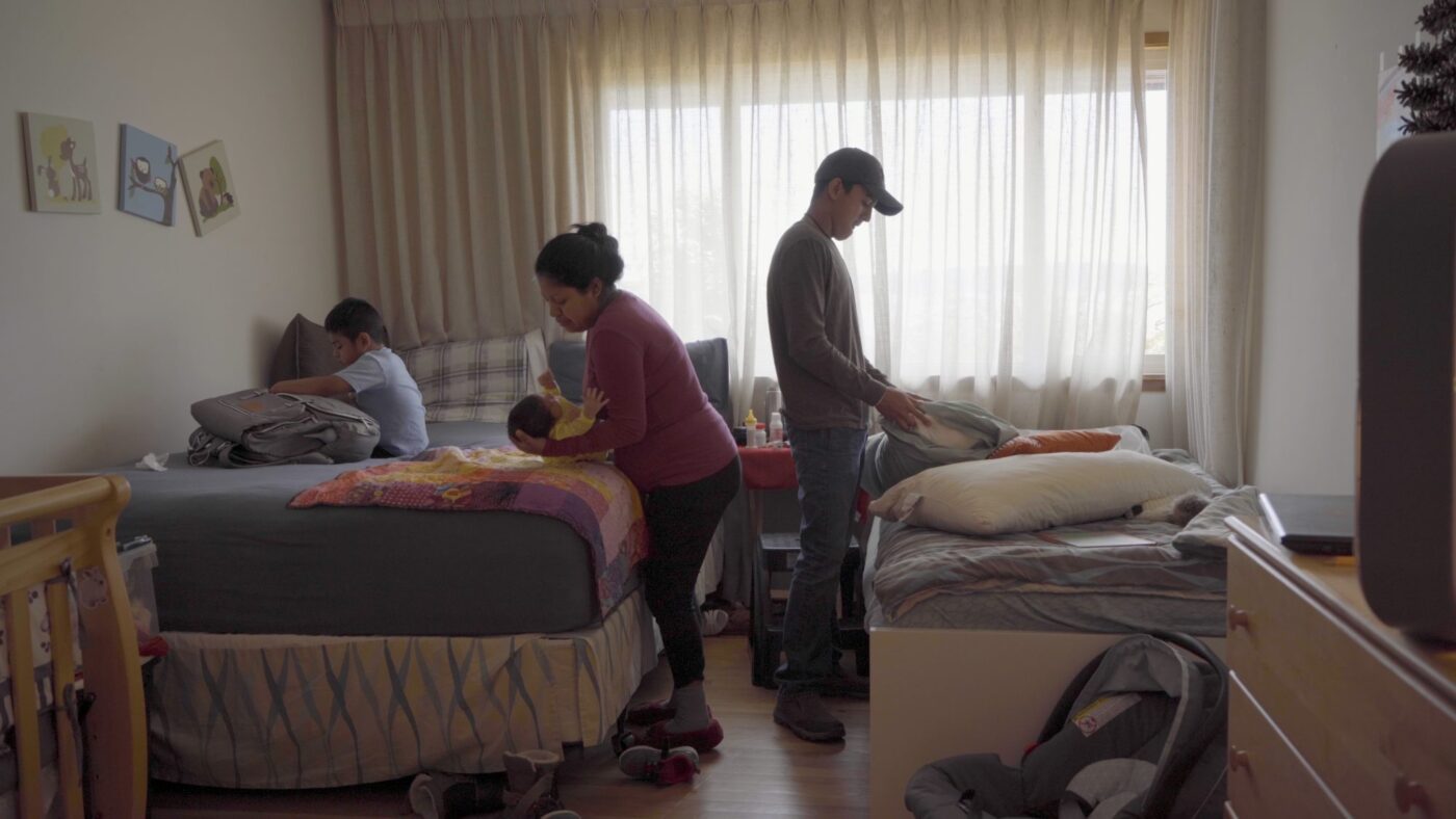 A family is occupied with different activities in a bedroom. One individual is comfortably lounging on the bed, engaged with their laptop, while the other two members are busy organizing the room.