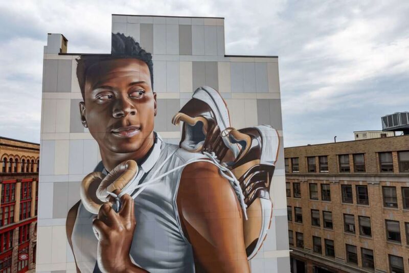 A huge wall painting showcasing an individual holding a pair of sneakers on a building side.