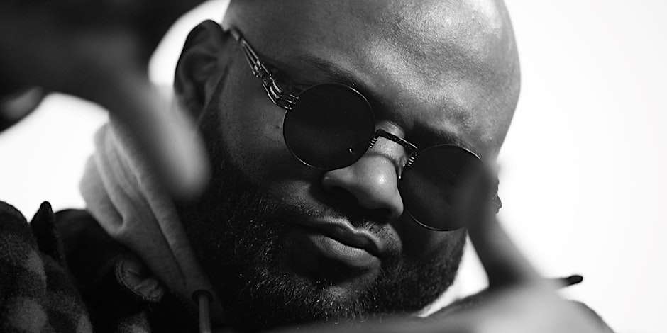 The content features a black and white image of a stylish, bald man with round sunglasses, uniquely posing with his hands partially covering his face.