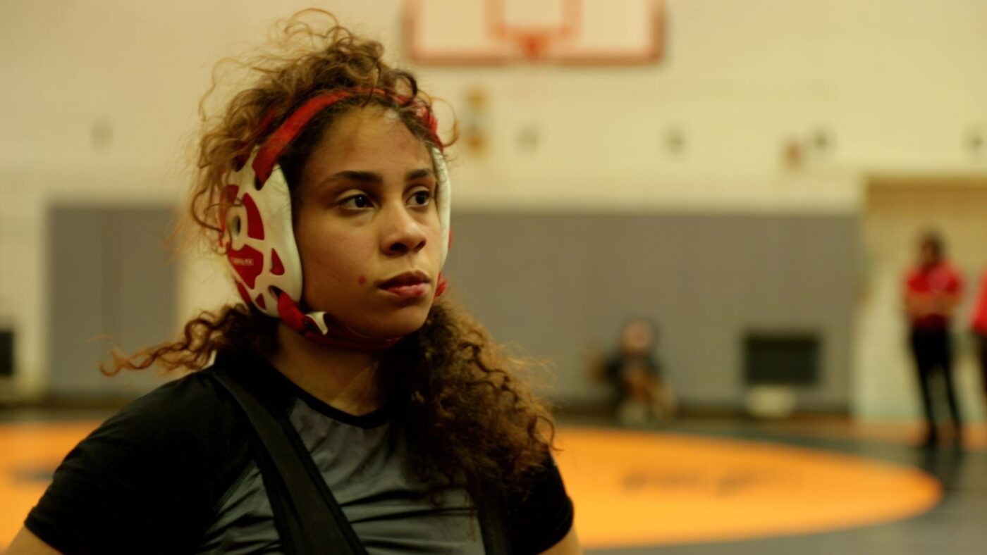 Female wrestler wearing protective headgear practicing in a gym.