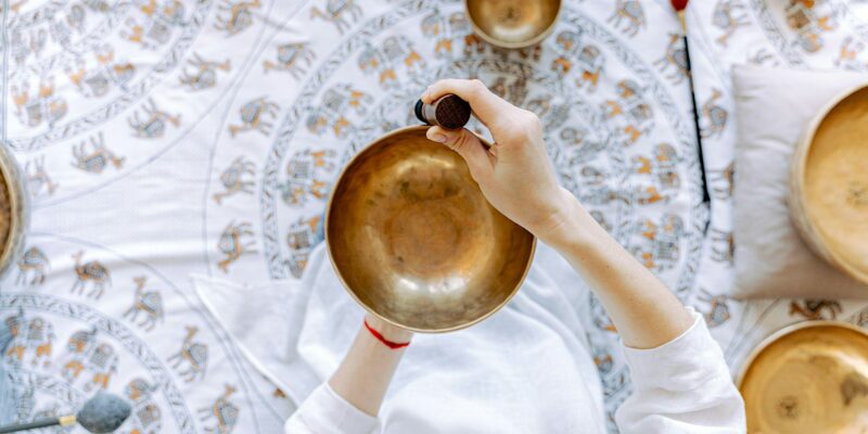 An individual is shown holding a petite singing bowl and a mallet, set amidst a textured fabric backdrop. There are additional singing bowls arranged around this one.