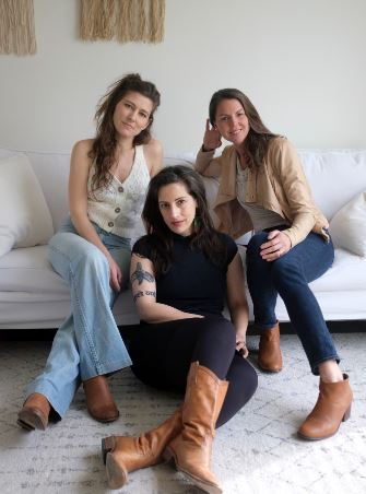 Three women striking a pose together in a space featuring a pristine white sofa and eye-catching wall decor in the backdrop.