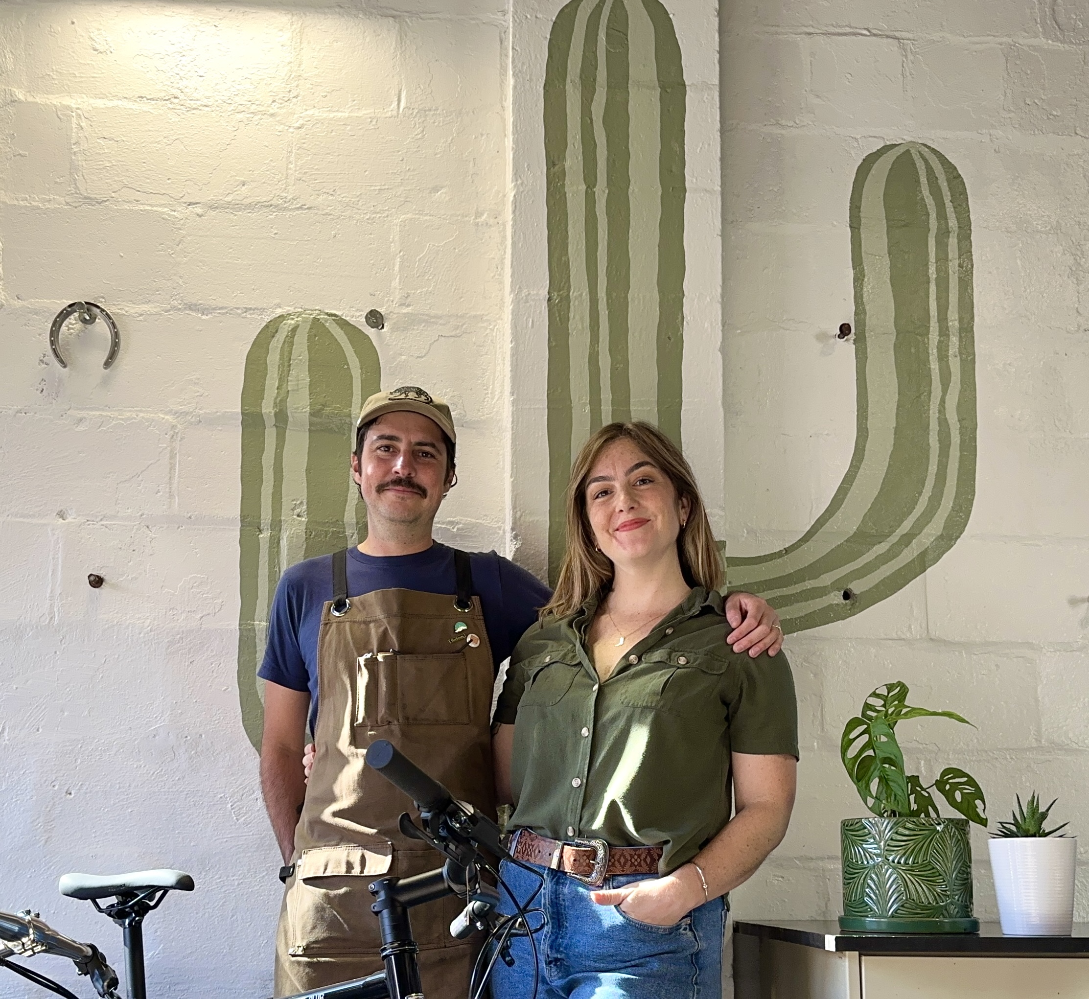 Introducing Our Team: Third Wheel - An inviting snapshot of two happy individuals, donning aprons while standing inside a café. A magnificent mural depicting a cactus serves as the backdrop to their joyous scene.