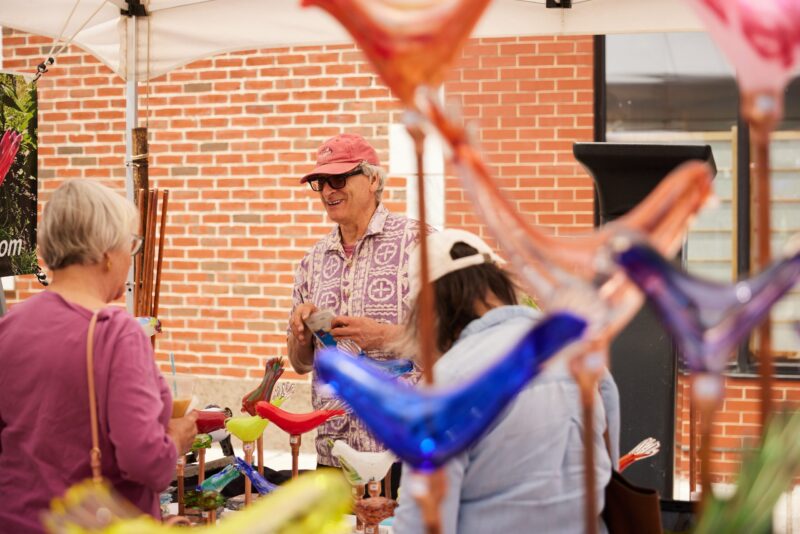 Elderly gentleman, donned in a pink hat and sweater, engaged in selling vibrant glass bird sculptures at a market booth while communicating with women shoppers.