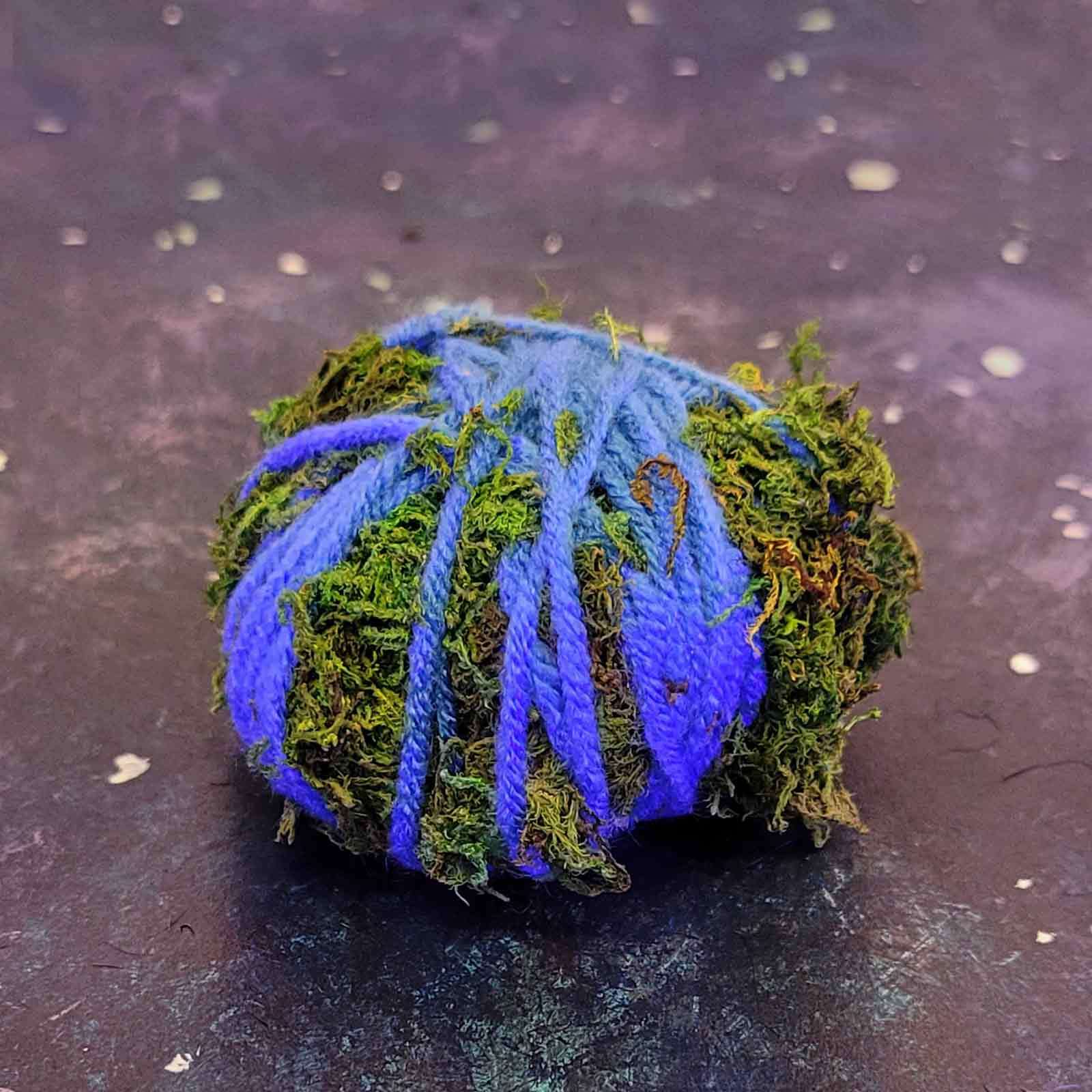 A green mossy item decorated with strands of blue yarn, set against a dappled dark backdrop.